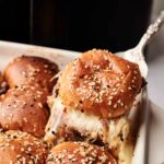 A serving spatula lifts a cheesy Air Fryer Roast Beef Slider with a seeded bun from a baking dish filled with similar sliders in front of a black kitchen appliance.