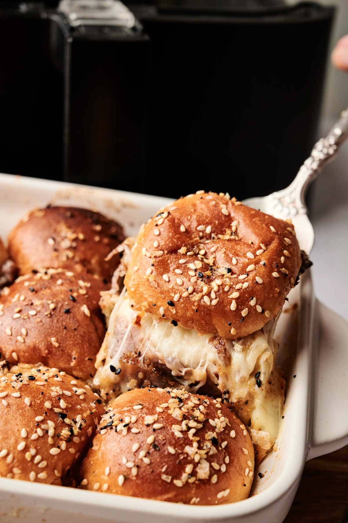 A serving spatula lifts a cheesy Air Fryer Roast Beef Slider with a seeded bun from a baking dish filled with similar sliders in front of a black kitchen appliance.