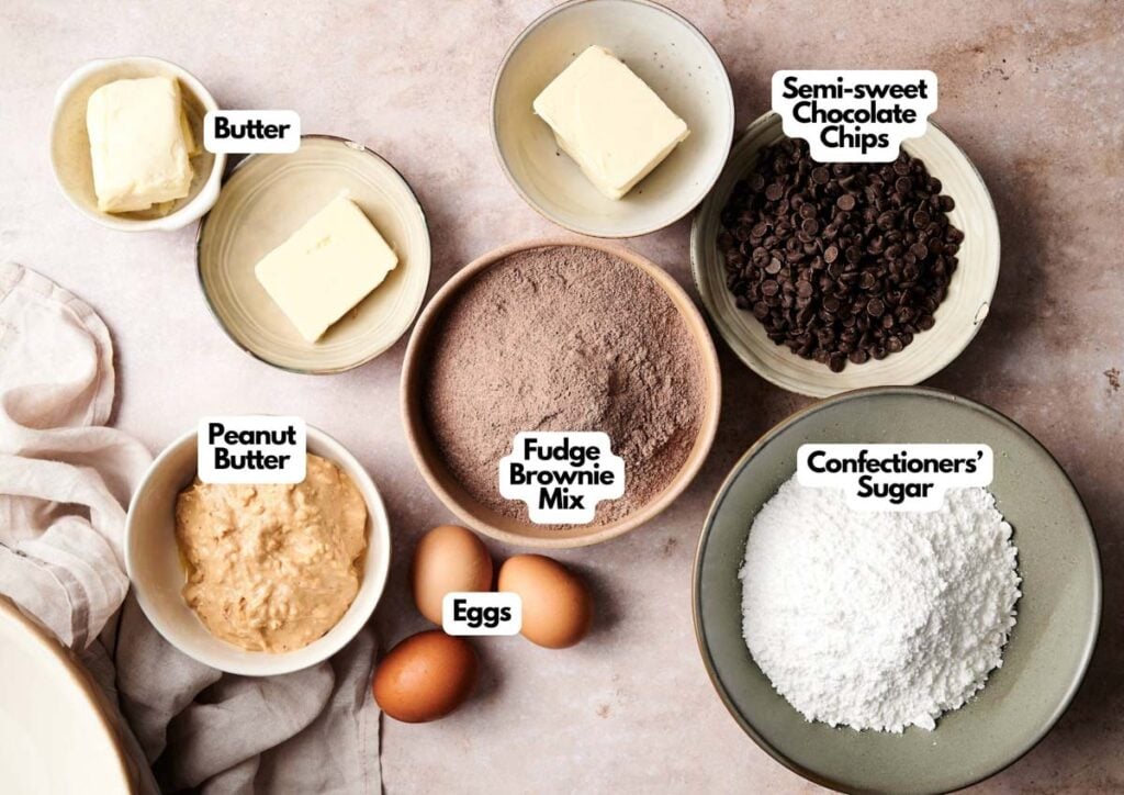 Bowls containing butter, peanut butter, eggs, fudge brownie mix, semi-sweet chocolate chips, and confectioners&rsquo; sugar are arranged on a countertop&mdash;perfect for preparing Air Fryer Buckeye Brownies. Each bowl is labeled with its ingredient.