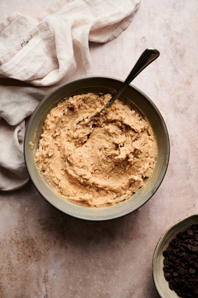 A bowl of peanut butter filling with a spoon inside, placed on a countertop next to a beige cloth and a small bowl of chocolate chips&mdash;perfect for whipping up Air Fryer Buckeye Brownies.