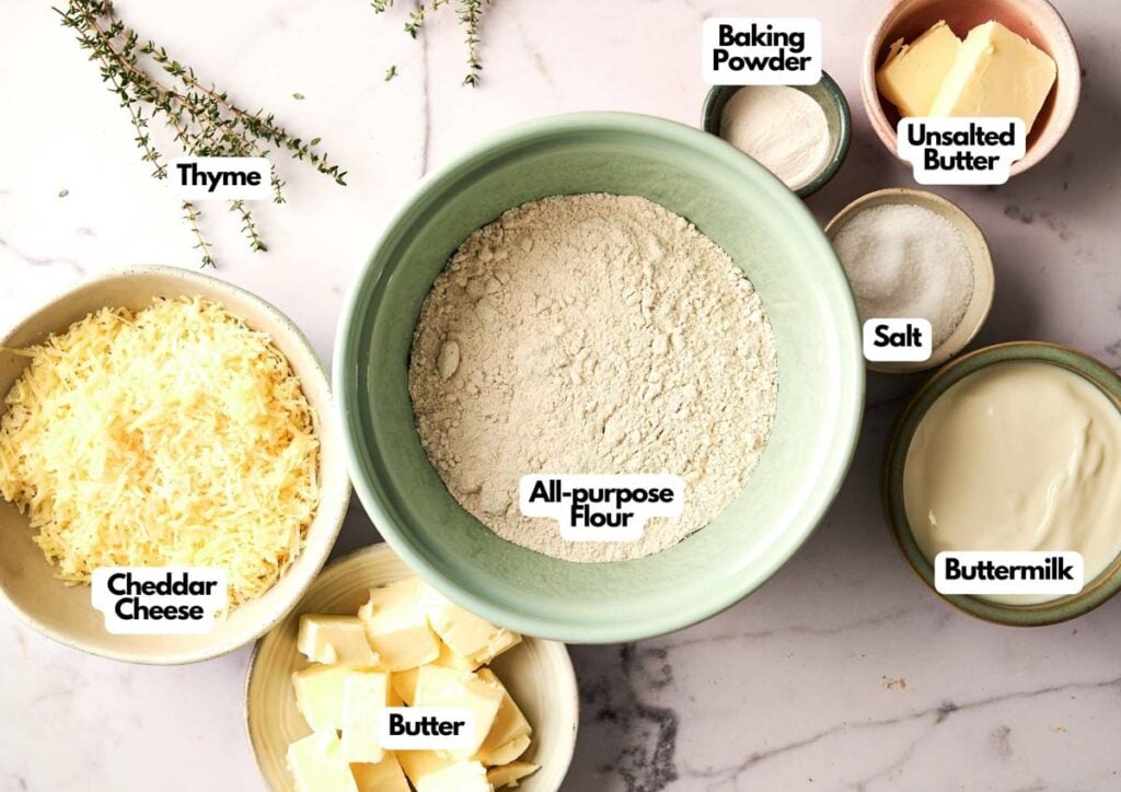 Assorted baking ingredients for Air Fryer Cheddar Biscuits in bowls labeled as thyme, cheddar cheese, butter, all-purpose flour, baking powder, unsalted butter, salt, and buttermilk on a marble surface.
