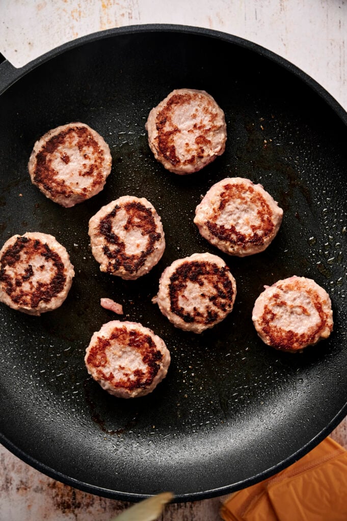 Seven browned sausage patties cooking in a black frying pan with a bit of oil, viewed from above&mdash;perfect as a savory side for Air Fryer Cheeseburger Sliders.