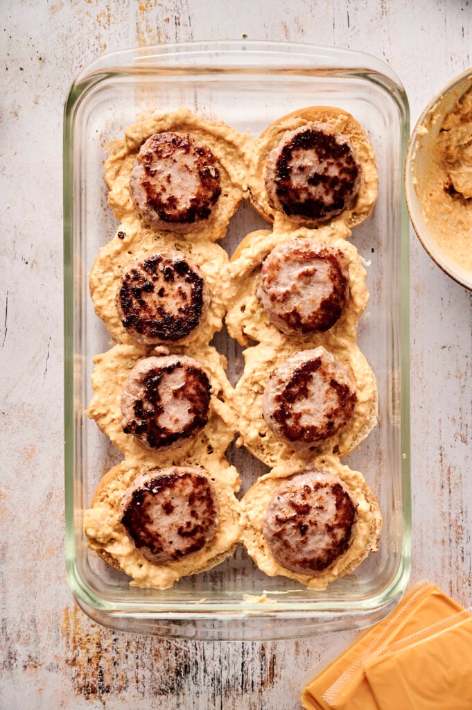 A glass baking dish with eight browned Air Fryer Cheeseburger Sliders nestled on scoops of a creamy cheese sauce, next to a bowl with more mixture and slices of processed cheese.