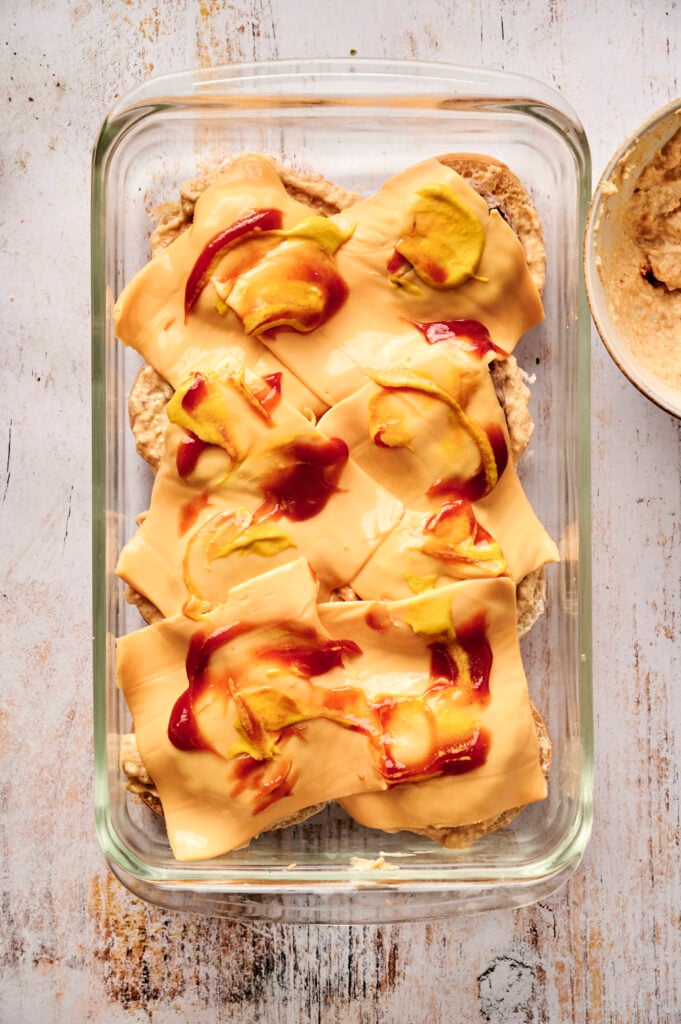 A glass baking dish containing layered Air Fryer Cheeseburger Sliders, with hamburger patties topped with sliced cheese, ketchup, and mustard, ready for baking.
