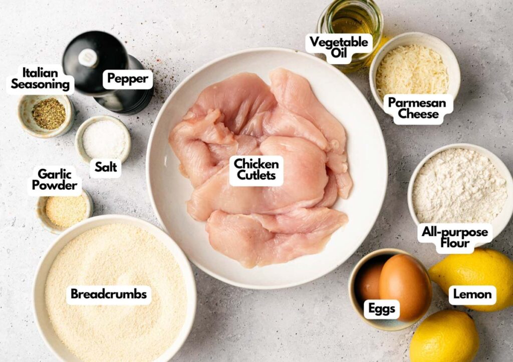 Overhead view of ingredients for Air Fryer Chicken Cutlets, including raw chicken, breadcrumbs, eggs, flour, parmesan, seasonings, salt, pepper, vegetable oil, garlic powder, and lemons.