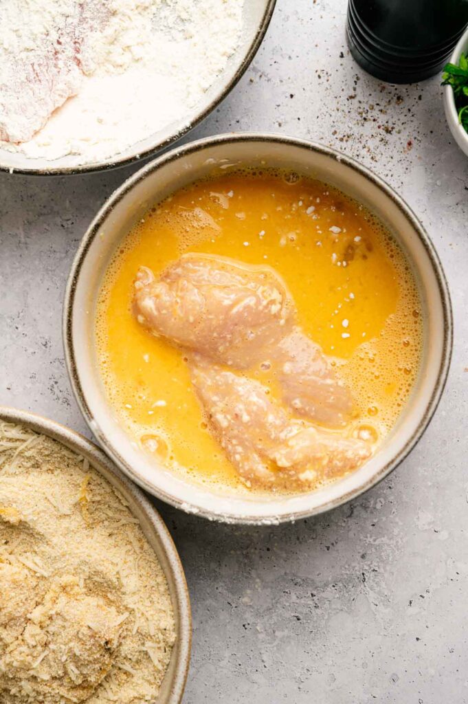 Raw chicken cutlets in a bowl of beaten eggs, with bowls of flour and breadcrumbs nearby on a gray countertop&mdash;perfect for making crispy Air Fryer Chicken Cutlets.
