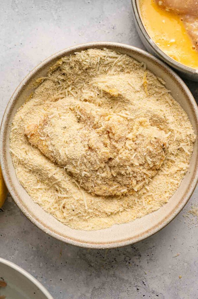 A piece of raw chicken is coated in breadcrumbs and grated cheese in a shallow bowl, with beaten eggs nearby&mdash;perfect preparation for making crispy Air Fryer Chicken Cutlets.