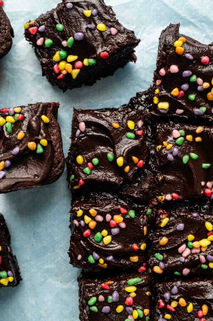 Nine Air Fryer Cosmic Brownies with chocolate frosting and colorful sprinkles are arranged on a light blue surface.