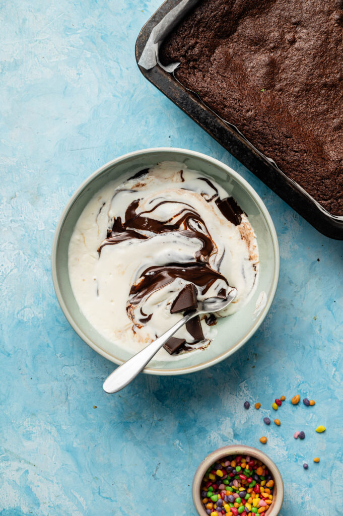 A bowl with vanilla ice cream and chocolate sauce, a spoon inside, sits next to a pan of Air Fryer Cosmic Brownies and a small bowl of colorful candy sprinkles on a blue surface.