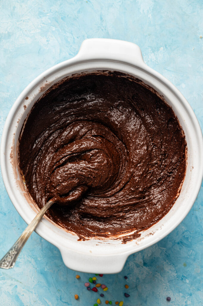 A white bowl filled with chocolate batter for Air Fryer Cosmic Brownies and a spoon, sitting on a blue surface with a few small colorful candies nearby.