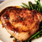 A cooked pork chop with a brown glaze, seasoned with herbs and reminiscent of Air Fryer Ham Steaks, served alongside steamed asparagus on a beige plate.