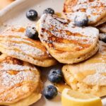 A plate of fluffy Air Fryer Lemon Ricotta Pancakes topped with powdered sugar, fresh blueberries, lemon slices, and drizzled with syrup.