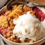 A bowl of Air Fryer Rhubarb Crisp topped with a scoop of melting vanilla ice cream, with a spoon resting in the bowl.
