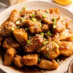 A plate of sesame chicken pieces topped with sesame seeds and chopped green onions, coated in a glossy brown sauce reminiscent of Panda Express Orange Chicken.