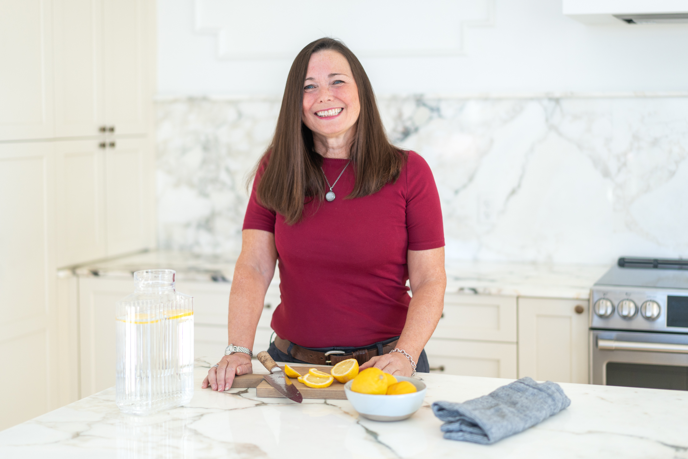 A woman stands in a modern kitchen, smiling, with sliced lemons and oranges on a cutting board, a bowl of lemons, a pitcher of lemon water, and a folded towel on the counter.