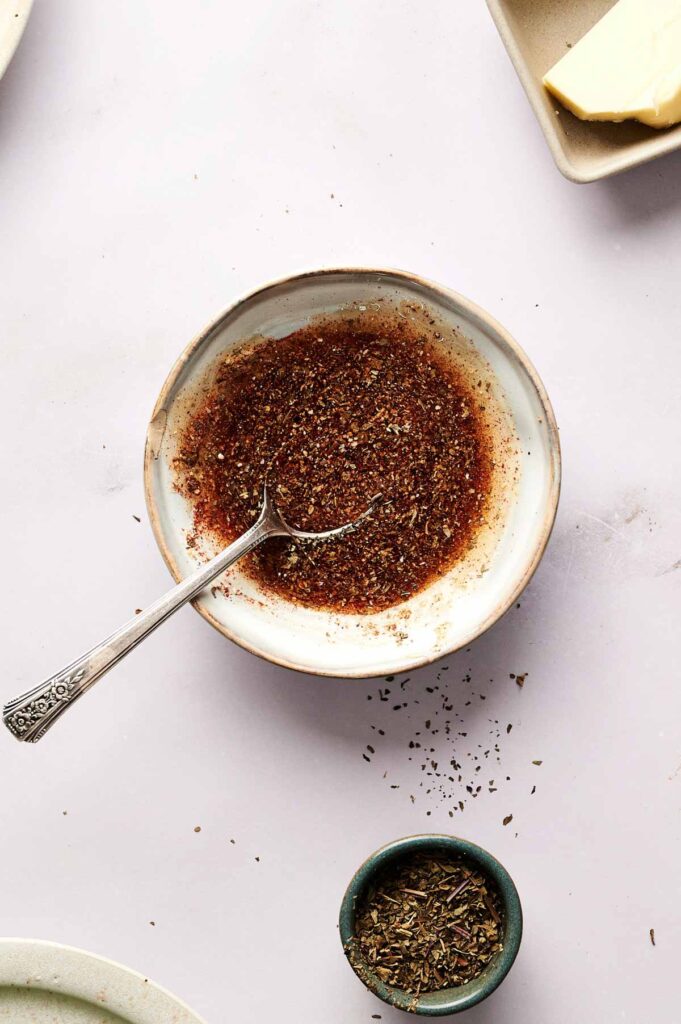 A bowl of ground spice mix with a spoon, some of the spice spilled on the table, and a small dish of dried herbs nearby—perfect for seasoning Air Fryer Ham Steaks to add an extra layer of flavor.