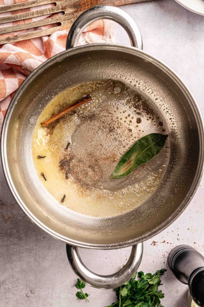 A stainless steel pot with bubbling butter, whole spices including a bay leaf, a cinnamon stick, and cloves—the perfect aromatic base for preparing Lamb Rogan Josh—sits on a light-colored countertop.