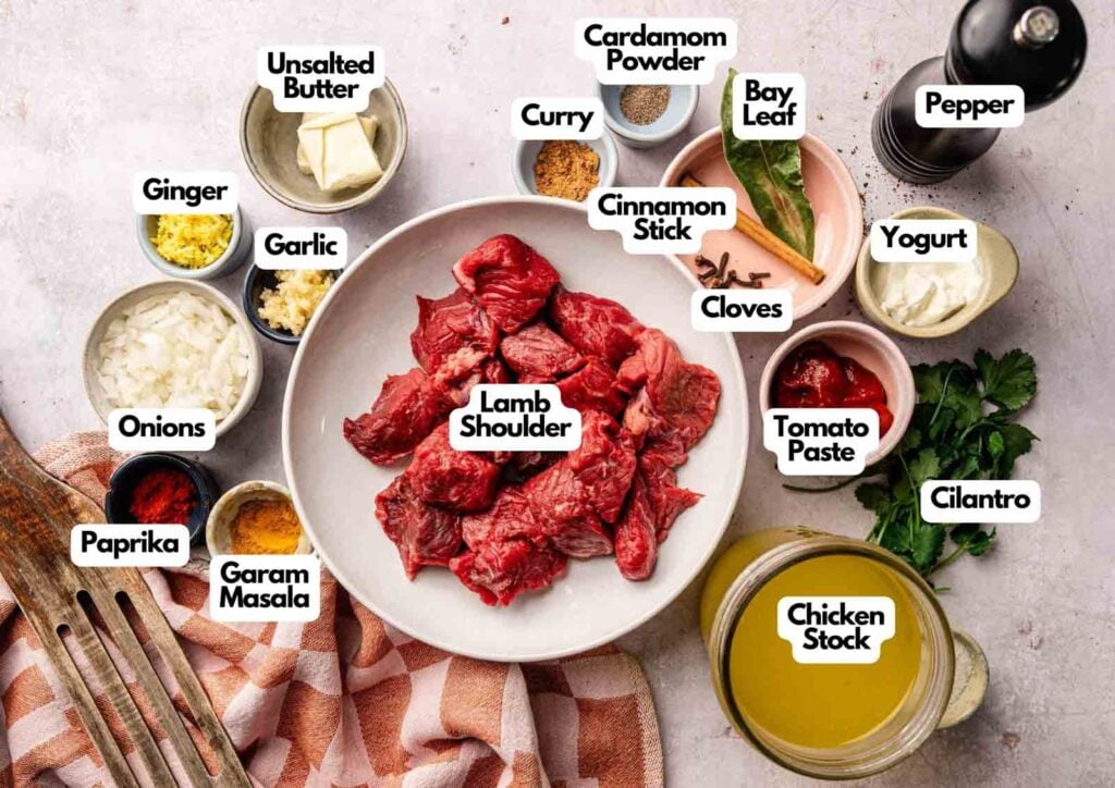 A plate of raw lamb shoulder, ideal for Lamb Rogan Josh, is surrounded by small bowls containing labeled spices, herbs, butter, yogurt, tomato paste, chicken stock, onions, and garlic.