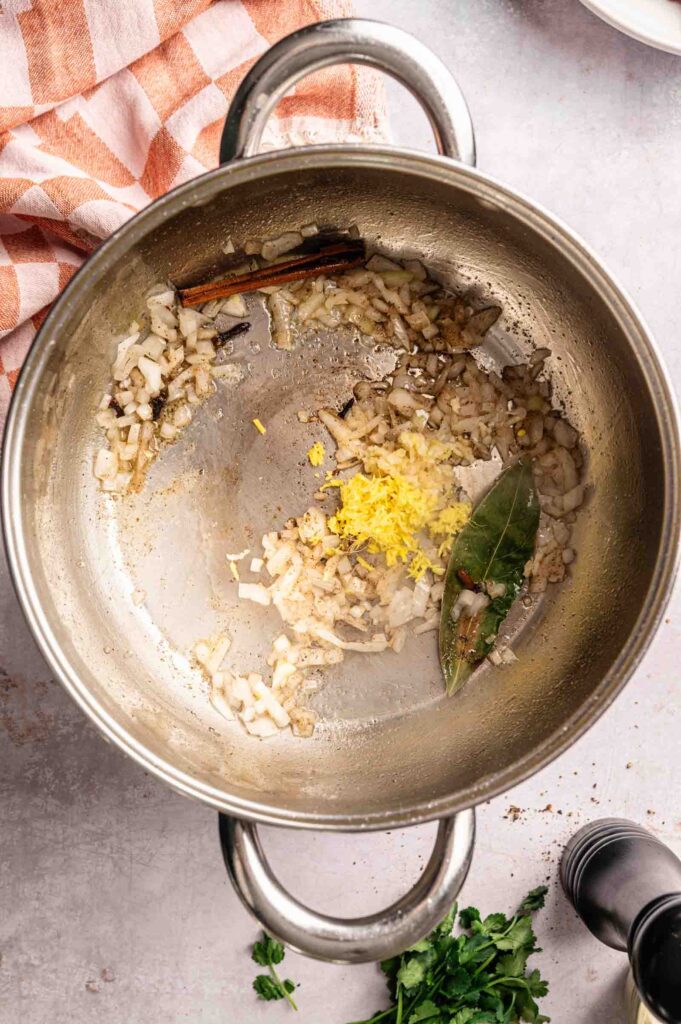 Chopped onions, grated ginger, a bay leaf, and a cinnamon stick sauté in a large metal pot next to a checked towel and some fresh herbs—essential aromatics forming the flavorful base for Lamb Rogan Josh.