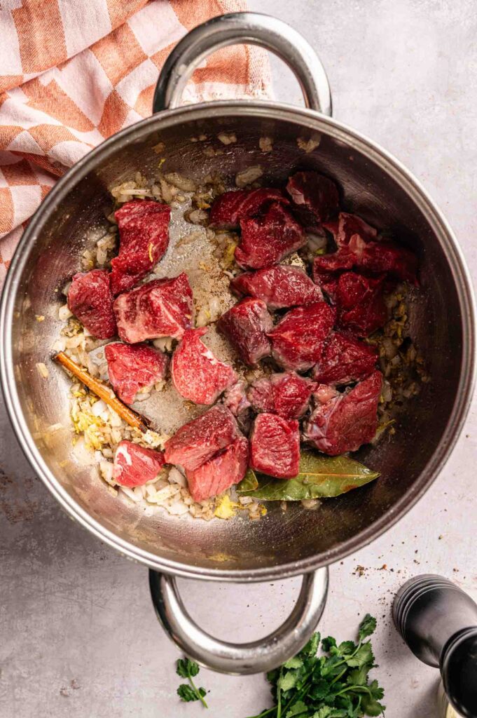 Chunks of raw beef, onions, bay leaf, and a cinnamon stick are arranged in a metal pot, ready for cooking—a preparation reminiscent of classic Lamb Rogan Josh. A striped cloth and some parsley are nearby.