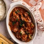 A white oval dish filled with Lamb Rogan Josh in a rich red sauce, garnished with chopped herbs, sits next to a bowl of rice, a wooden cutting board, and a checked cloth napkin.