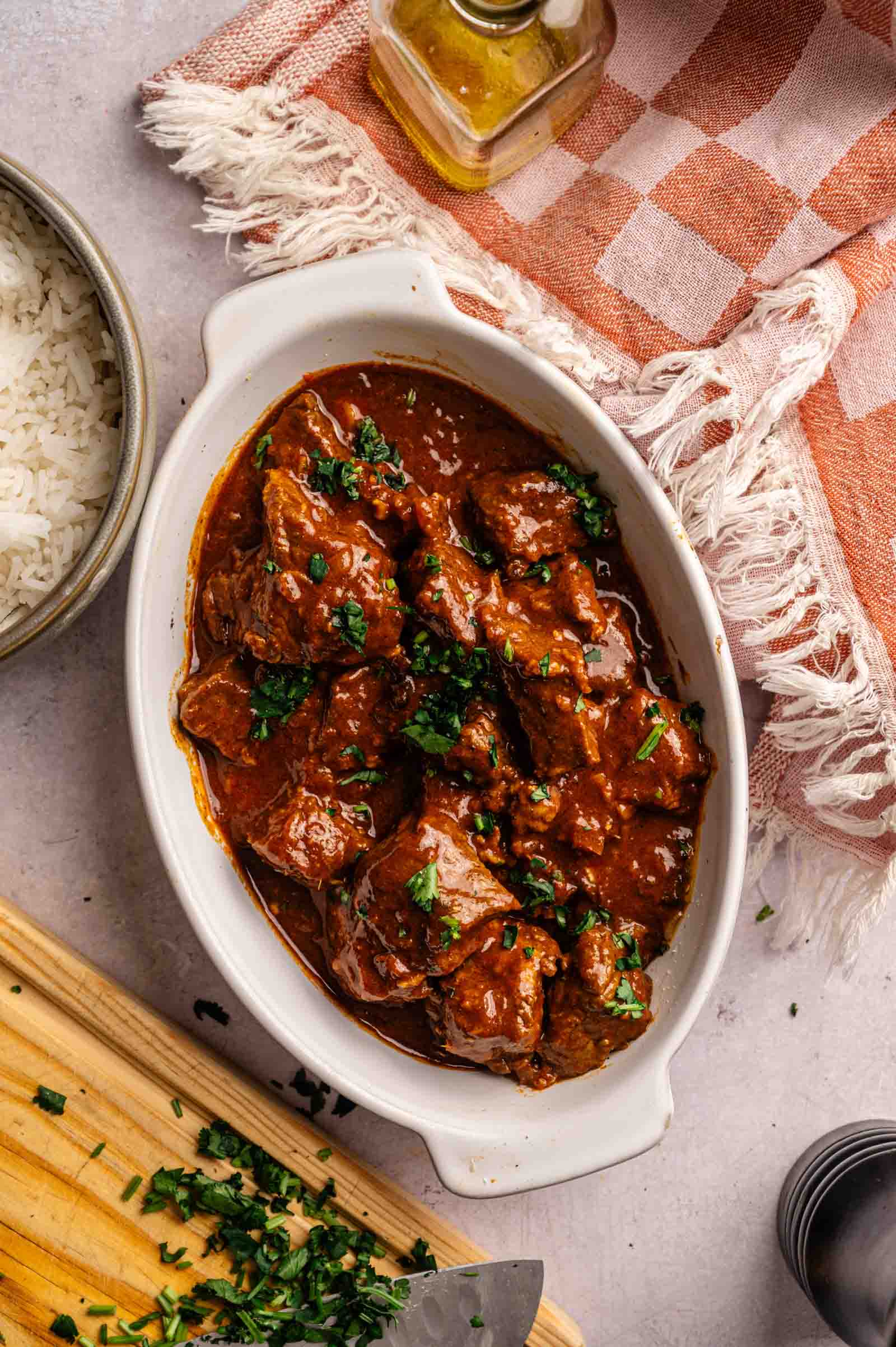 A white oval dish filled with Lamb Rogan Josh in a rich red sauce, garnished with chopped herbs, sits next to a bowl of rice, a wooden cutting board, and a checked cloth napkin.