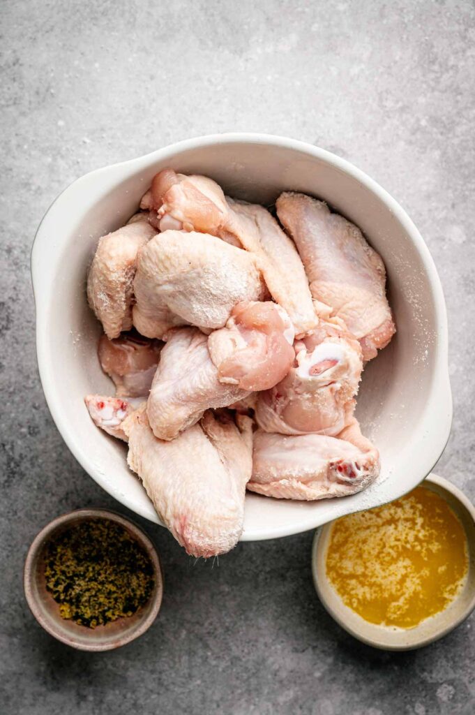 Raw chicken wings in a white bowl, with a small bowl of seasoning and a small bowl of melted butter on a gray surface—perfect ingredients for making delicious Air Fryer Lemon Pepper Wings.