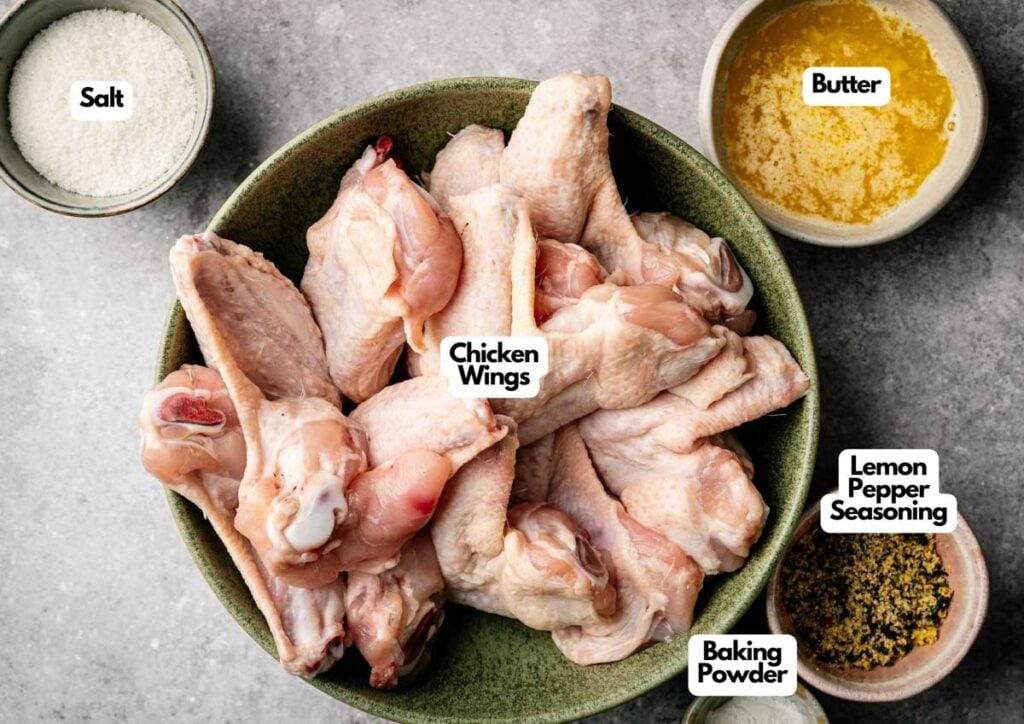 A bowl of raw chicken wings, perfect for making Air Fryer Lemon Pepper Wings, is surrounded by small bowls of salt, melted butter, lemon pepper seasoning, and baking powder on a gray surface.