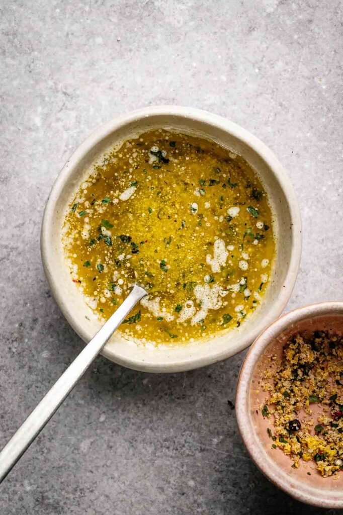 A bowl of seasoned olive oil with herbs and spices, perfect for drizzling over Air Fryer Lemon Pepper Wings, sits with a spoon inside. Another small bowl with herb mixture rests nearby on a gray surface.