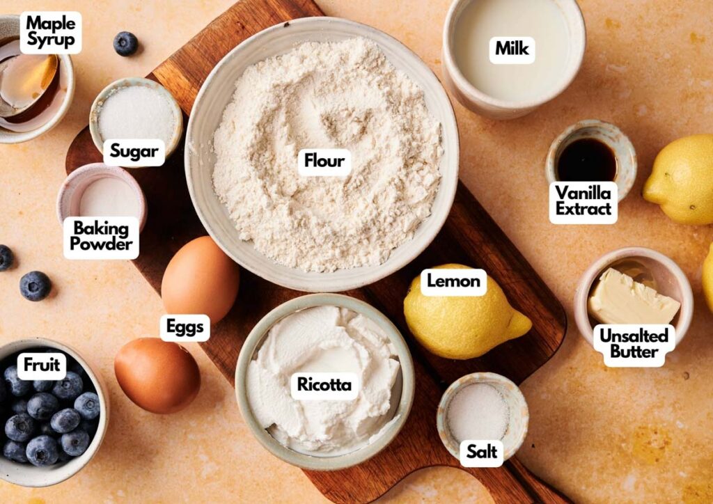 Various baking ingredients for Air Fryer Lemon Ricotta Pancakes&mdash;including flour, sugar, eggs, ricotta, lemon, and blueberries&mdash;are arranged on a countertop.