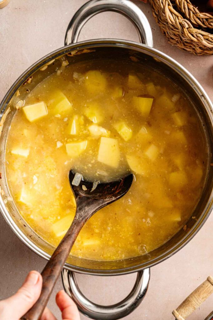A pot of Loaded Baked Potato Soup simmers as diced potatoes cook in broth, gently stirred with a wooden spoon.