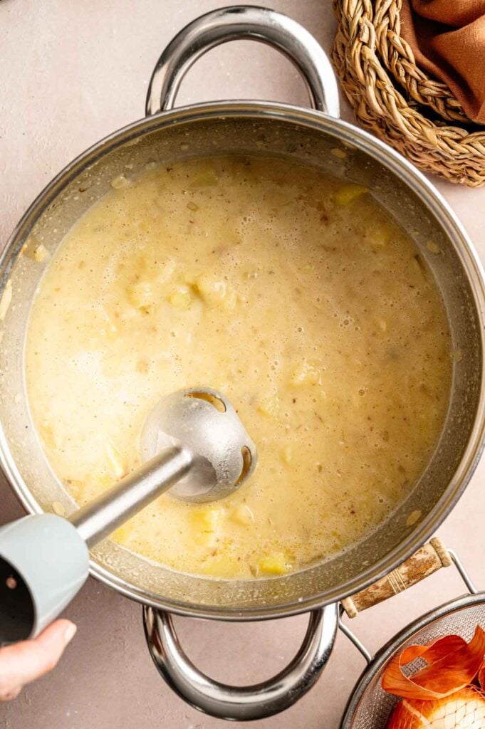 A large pot of creamy loaded baked potato soup is being blended with an immersion blender. A basket of onions and a dish towel are partially visible nearby.