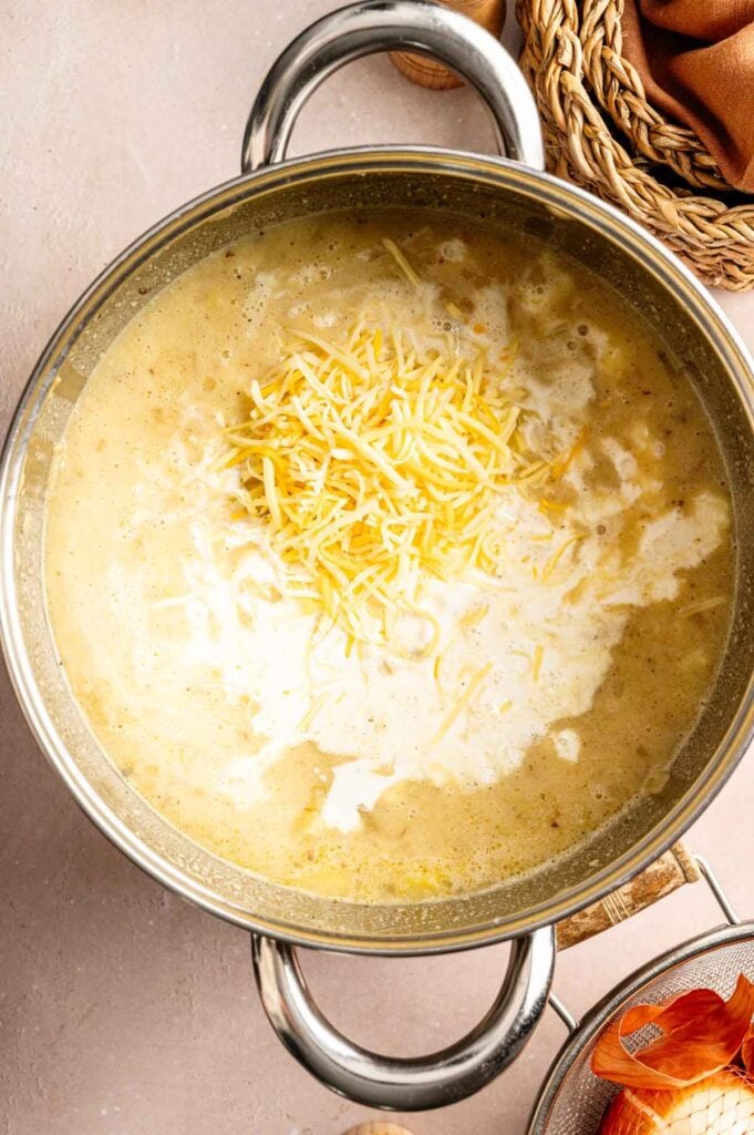 A pot of creamy Loaded Baked Potato Soup topped with shredded cheese, shown from above on a light countertop next to an onion and a woven basket.