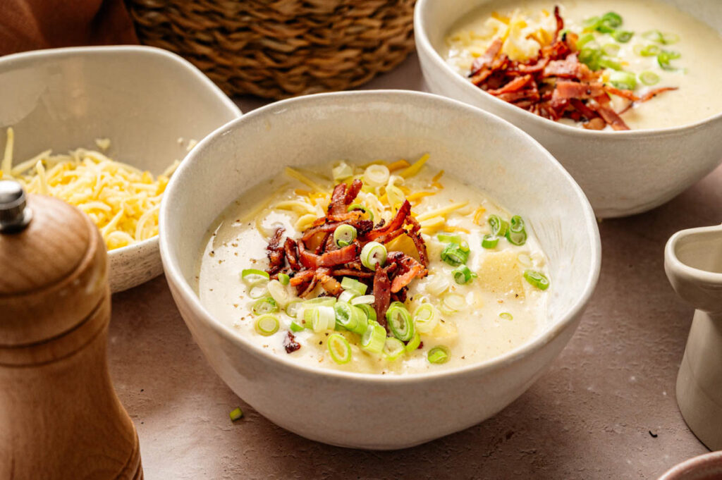 Two bowls of creamy Loaded Baked Potato Soup topped with shredded cheese, bacon, and sliced green onions, with a pepper grinder and cheese bowl nearby.