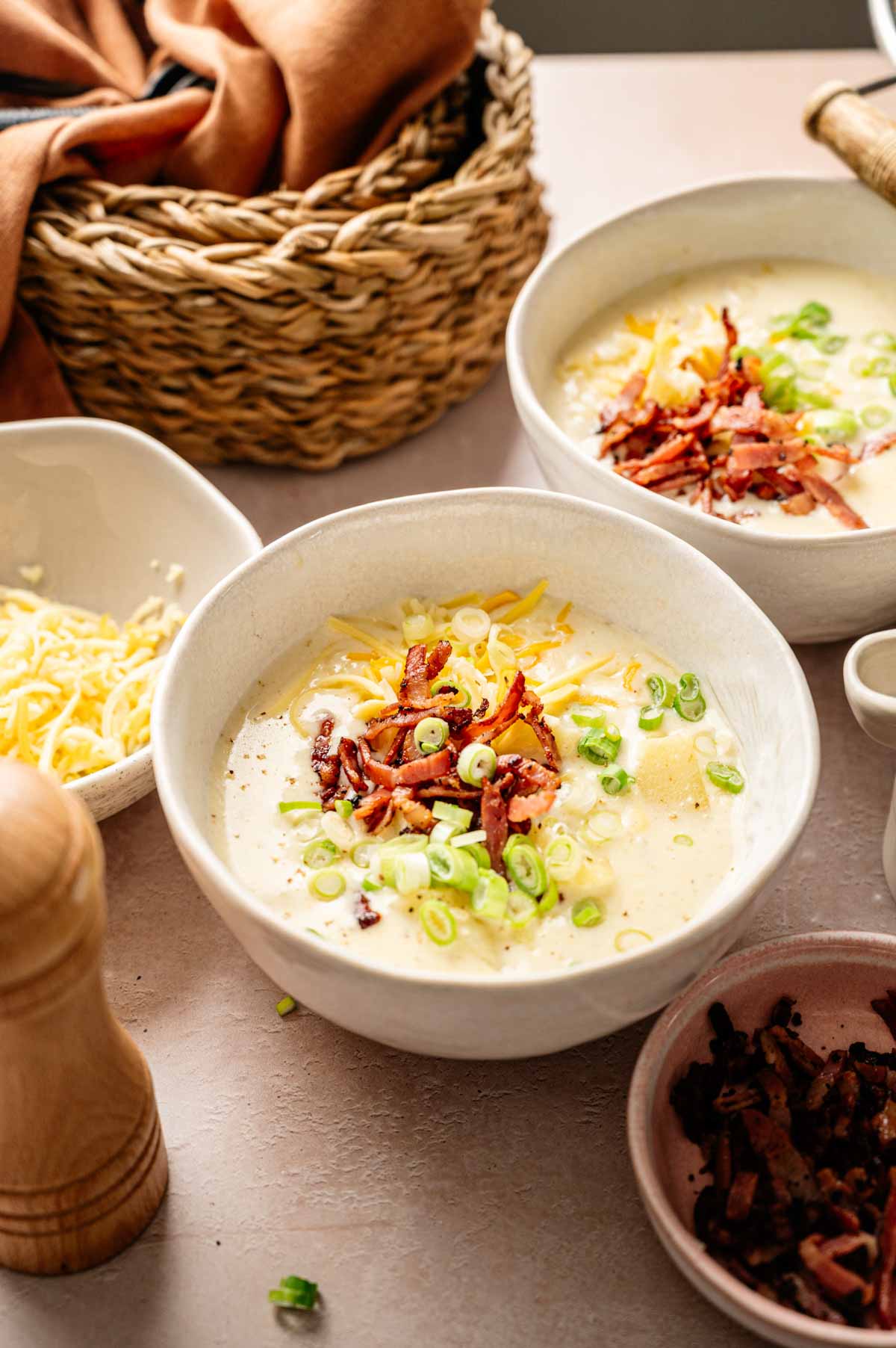 Two bowls of creamy Loaded Baked Potato Soup topped with shredded cheese, chopped green onions, and crispy bacon bits on a table with extra grated cheese and bacon on the side.