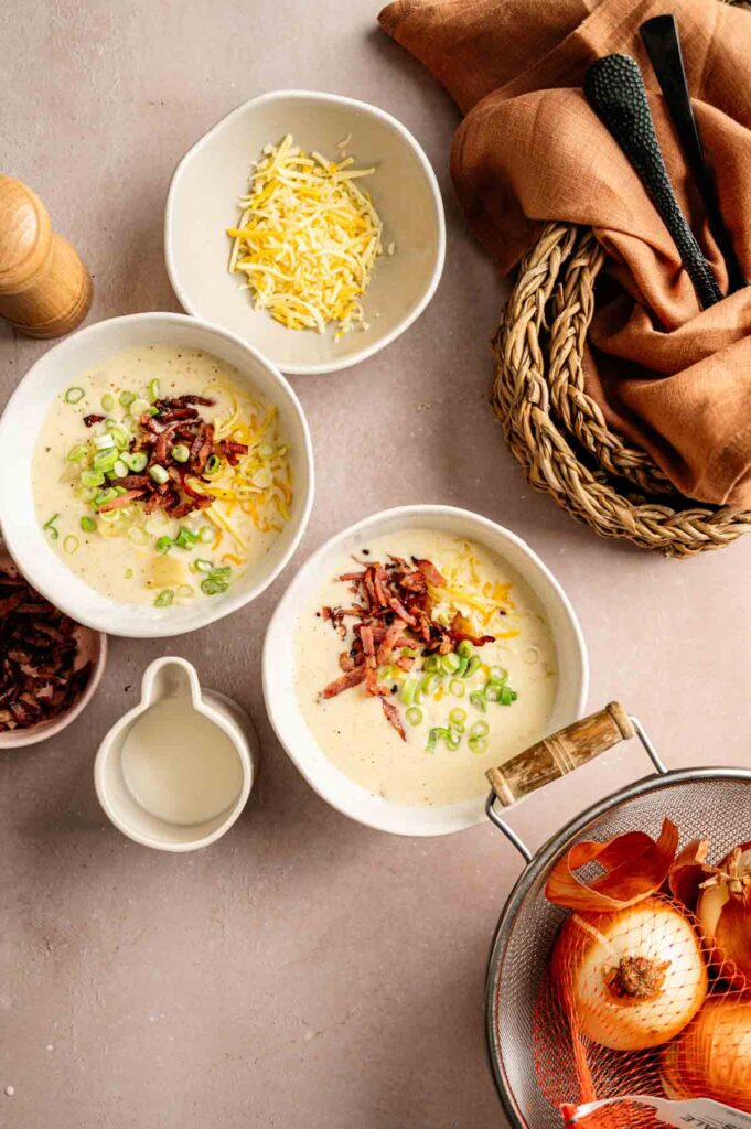 Two bowls of Loaded Baked Potato Soup topped with shredded cheese, bacon bits, and chopped green onions, with ingredients and utensils arranged around them on a countertop.