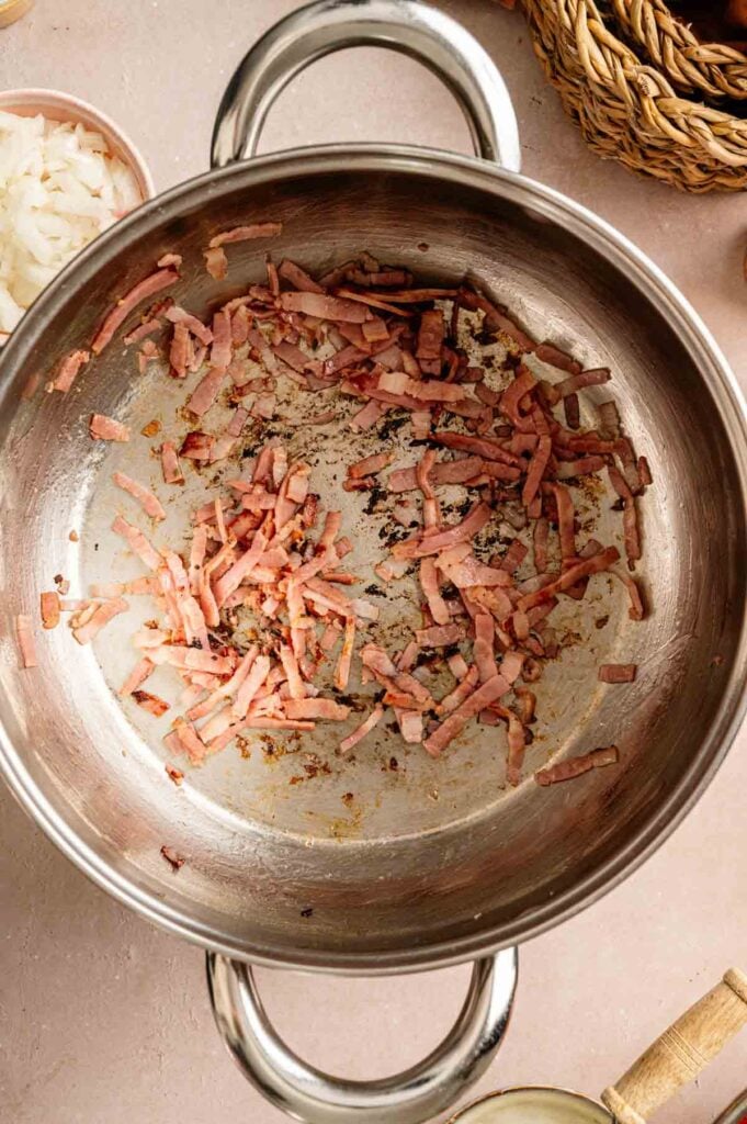 Chopped ham strips are sautéing in a large stainless steel pot, showing browning and fond at the bottom—a savory base for Loaded Baked Potato Soup as diced onions wait in a bowl nearby.