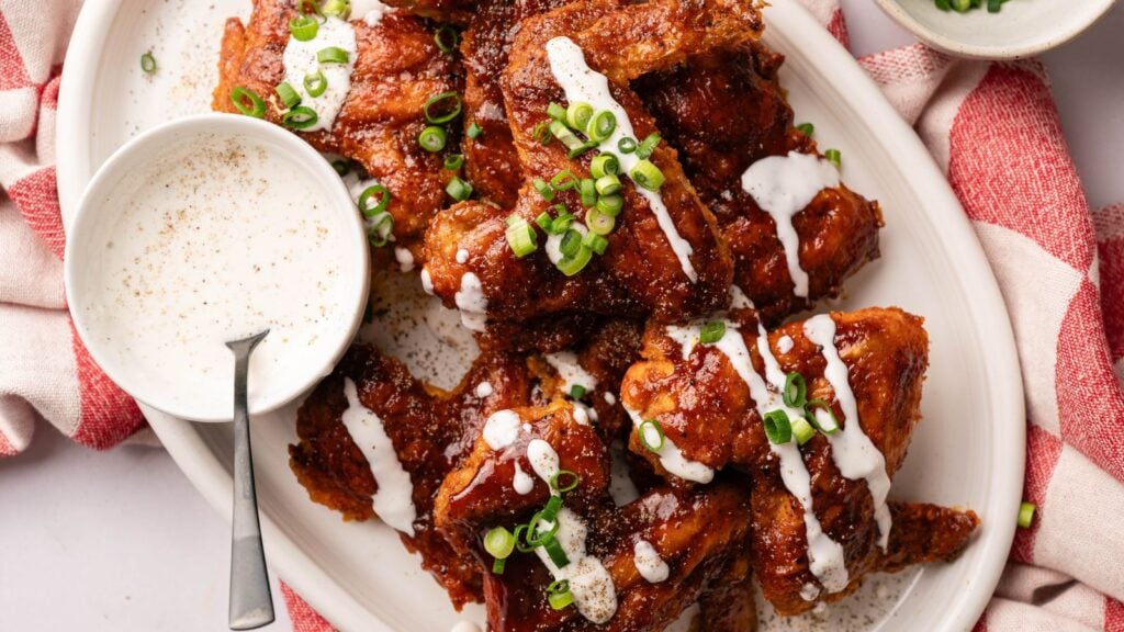 A plate of saucy chicken wings garnished with sliced green onions and drizzled with ranch dressing, served with a bowl of dipping sauce and a spoon.