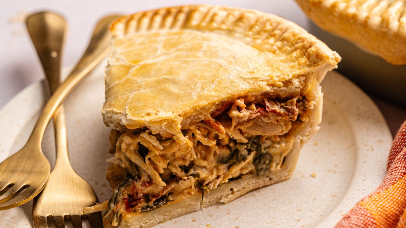 A slice of savory pie with a golden crust, filled with shredded chicken and vegetables, sits on a plate next to a gold fork and knife.
