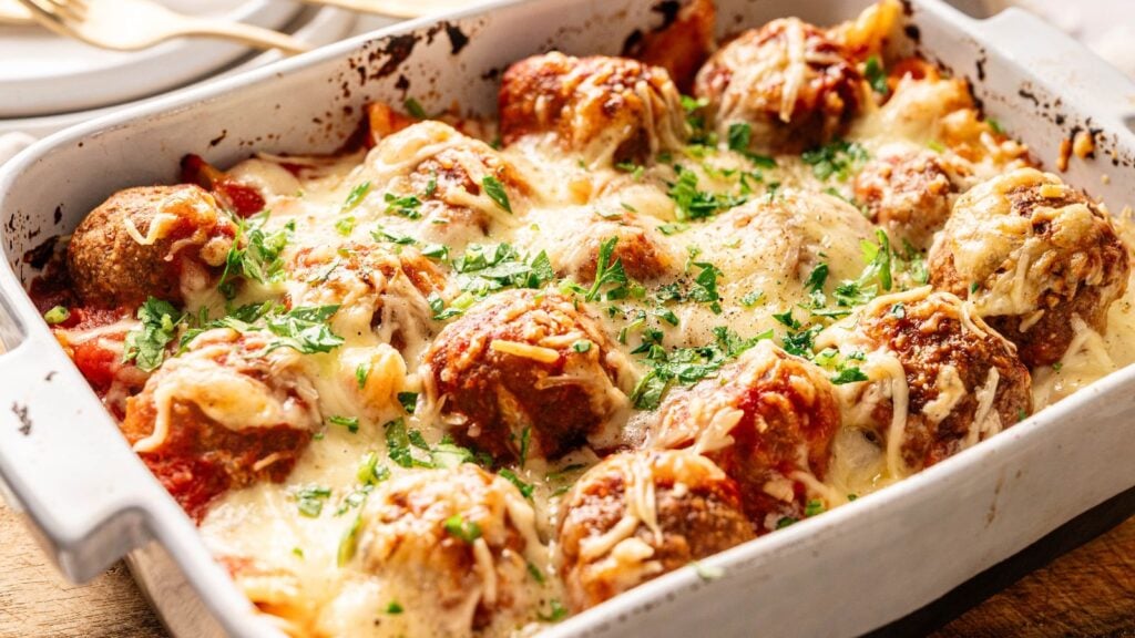 A baked casserole dish filled with meatballs topped with melted cheese, tomato sauce, and chopped herbs.