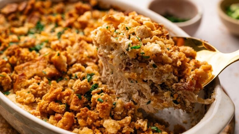 A close-up of a baked casserole with a golden, crumbly topping being served with a large spoon from a white baking dish.