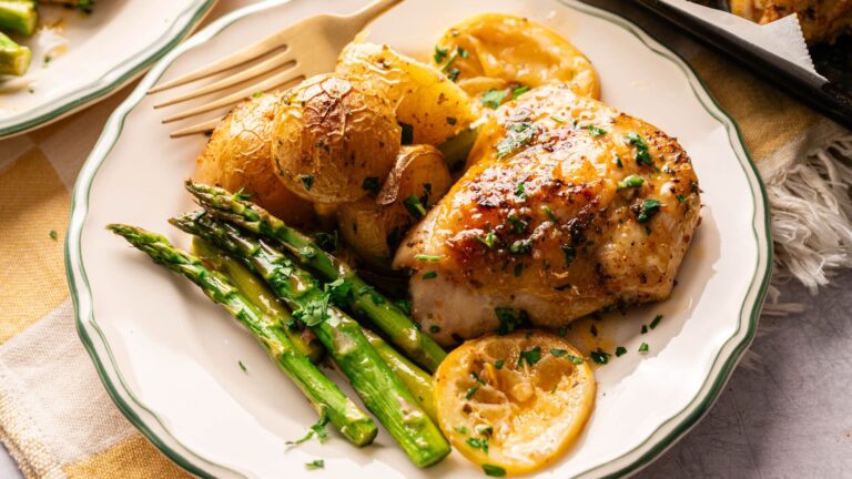 A plate with roasted chicken breast, lemon slices, asparagus spears, and golden roasted potatoes, garnished with chopped herbs.