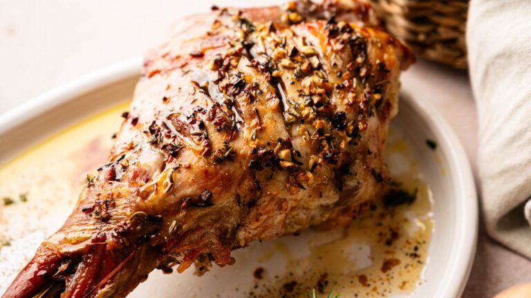 A roasted leg of lamb garnished with herbs and spices sits on a white plate, with juices pooling at the bottom.