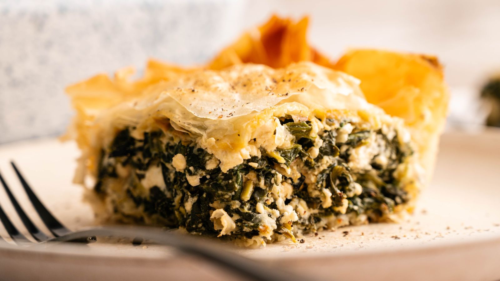 A slice of spinach and cheese pie with flaky pastry crust on a plate, next to a fork.