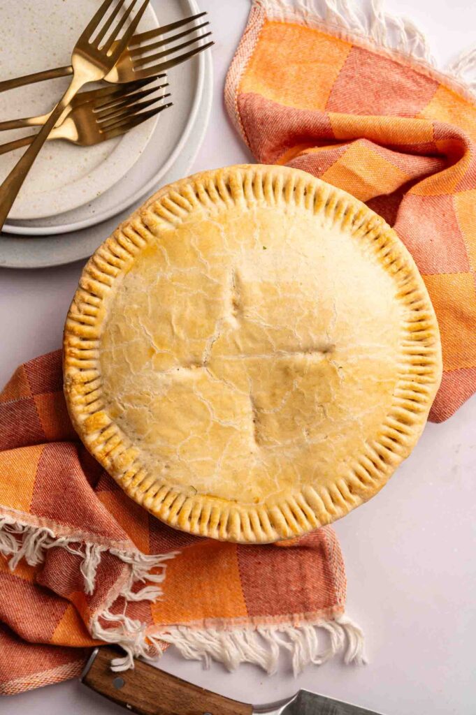 A Marry Me Chicken Pot Pie with a golden crust sits on an orange checkered cloth, next to plates, gold forks, and a pastry server.