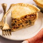 A slice of savory Marry Me Chicken Pot Pie filled with shredded chicken and vegetables sits on a plate with a fork and knife, next to an orange cloth napkin.