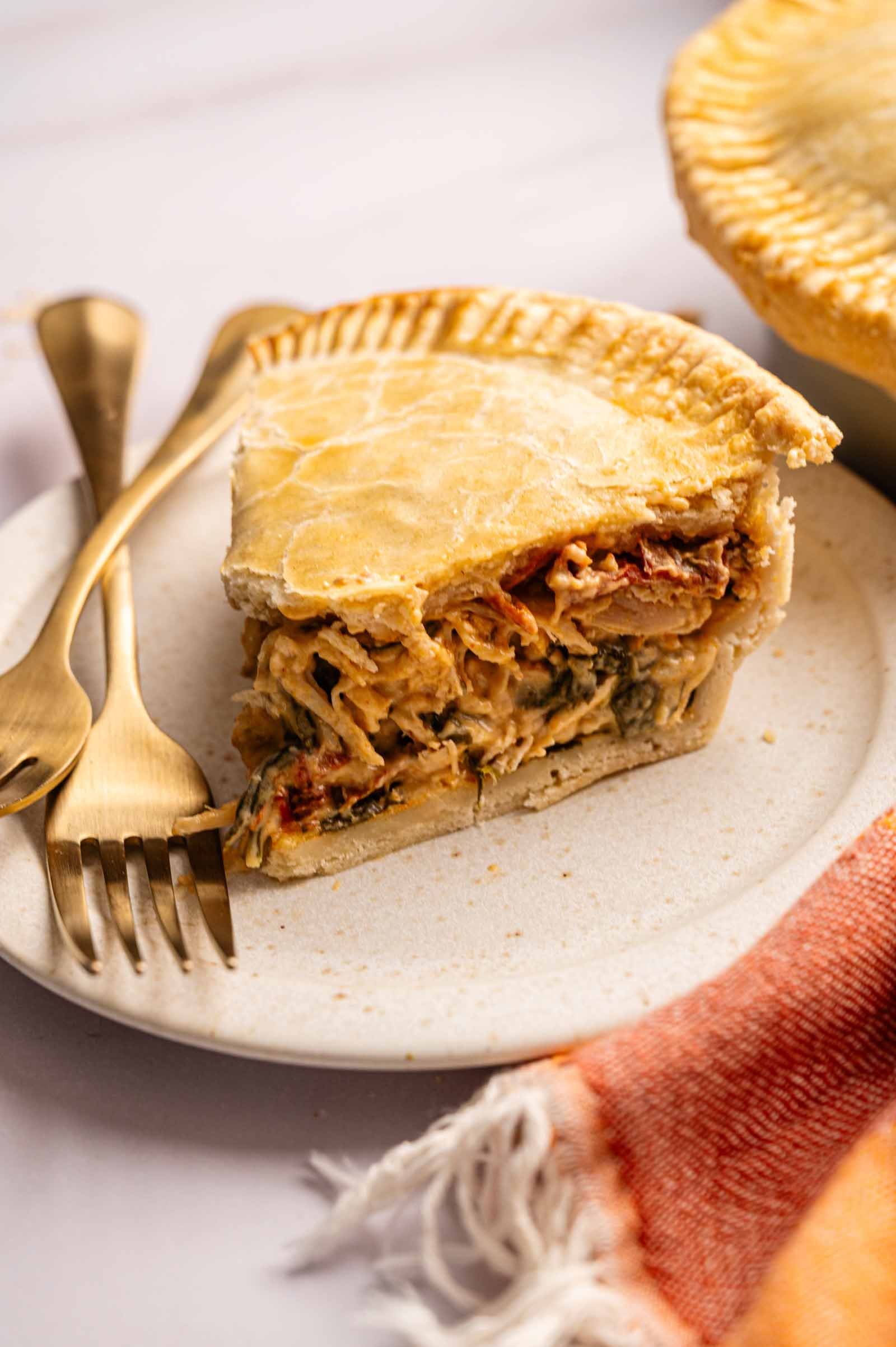 A slice of savory Marry Me Chicken Pot Pie filled with shredded chicken and vegetables sits on a plate with a fork and knife, next to an orange cloth napkin.