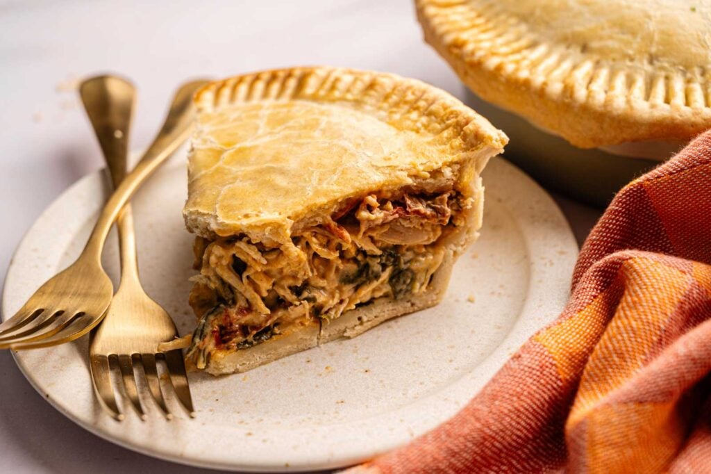 A slice of savory Marry Me Chicken Pot Pie with a golden crust is served on a plate with gold utensils, next to a whole pie and an orange napkin.
