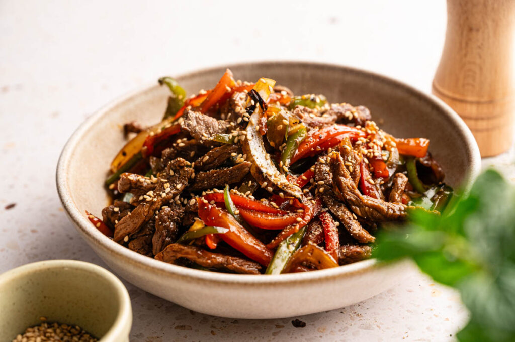 A bowl of stir-fried beef strips with red and green bell peppers, topped with sesame seeds, inspired by Panda Express Black Pepper Sirloin Steak.