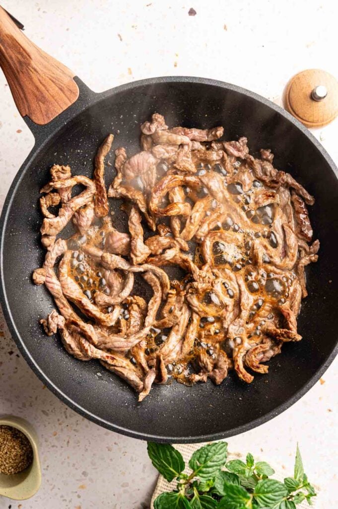 Thin strips of beef searing until browned in a black skillet.